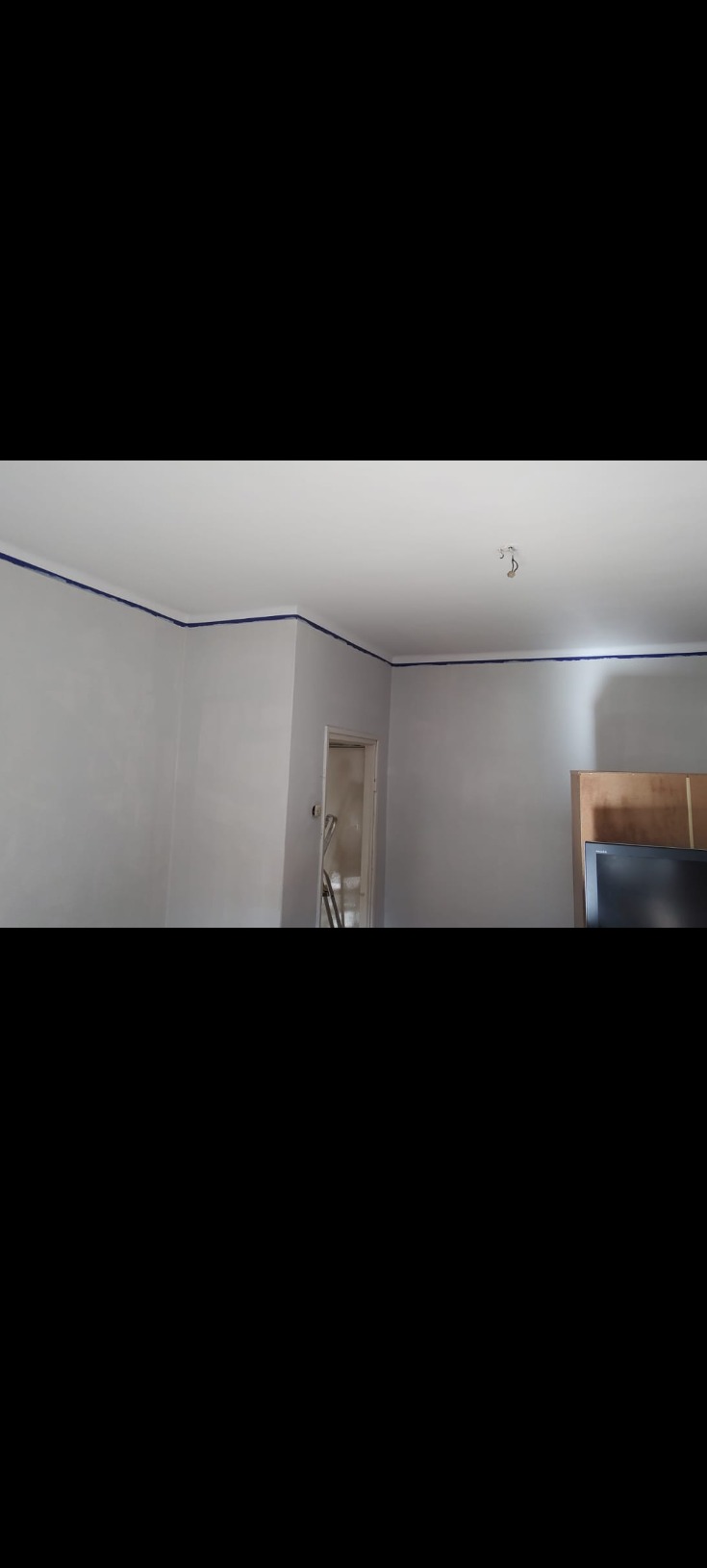 Świeżo pomalowane jasnoszare ściany z niebieską taśmą malarską przy suficie, przygotowane do dalszych prac, w tle widoczny fragment telewizora i drabina w przejściu.