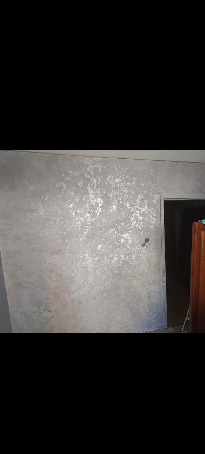 Ściana pokryta dekoracyjną szarą farbą z efektem srebrnych plam, widoczny fragment futryny drzwi z wystającym fragmentem mebla.