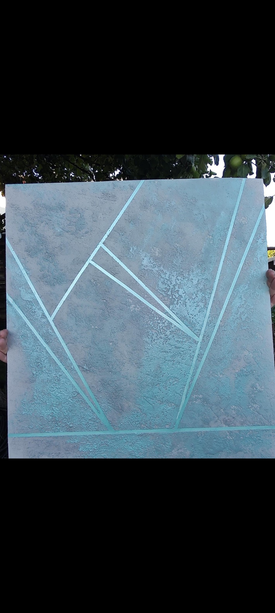 Dekoracyjna ściana z szarą strukturą i turkusowymi geometrycznymi liniami, prezentowana na tle zieleni drzew.