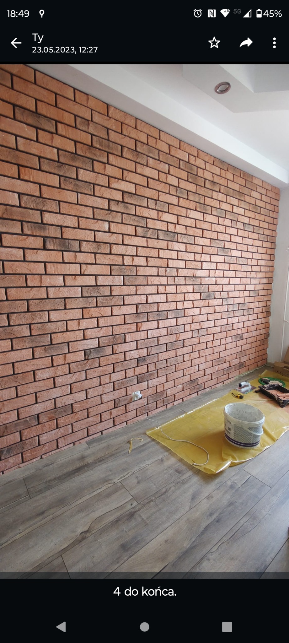 Ściana wyłożona dekoracyjną cegłą w odcieniach czerwieni i brązu, widoczna podczas prac wykończeniowych, z folią ochronną i wiadrem z farbą na podłodze z paneli.