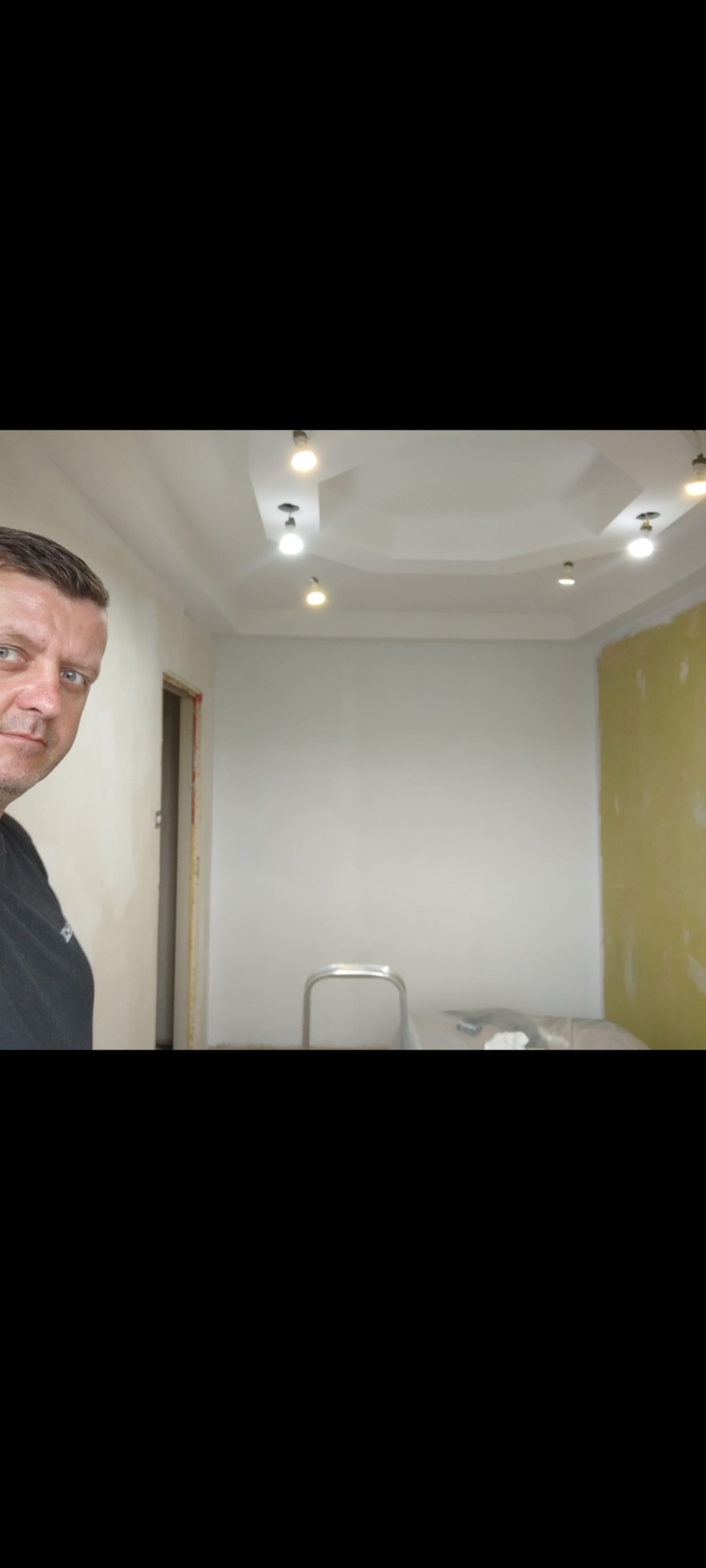 Pokój w trakcie malowania, widoczny pracownik z boku, częściowo pomalowane ściany, sufit z lampami, drabina w tle.