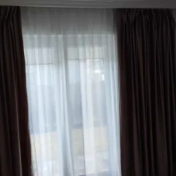 Okno z brązowymi zasłonami i delikatną białą firanką, widok na zewnątrz rozmyty przez światło.