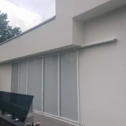 Szare, przesuwne osłony okienne na tarasie nowoczesnego domu z widocznym klimatyzatorem i szklaną balustradą.