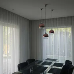 Elegancka jadalnia z dużymi oknami zasłoniętymi delikatnymi, białymi, plisowanymi zasłonami; szklany stół z czarnymi podkładkami i szklankami; trzy miedziano-srebrne lampy wiszące.