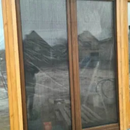 Brązowa rama okienna z zamontowaną siatką przeciw owadom, widok z zewnątrz na budynek w trakcie budowy.