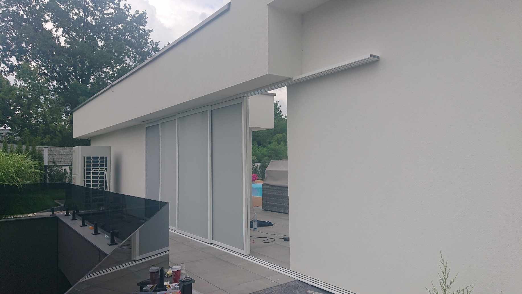 Zamknięte, przesuwne panele szklane w białej ramie, stanowiące osłonę tarasu nowoczesnego domu z widokiem na basen i ogród.