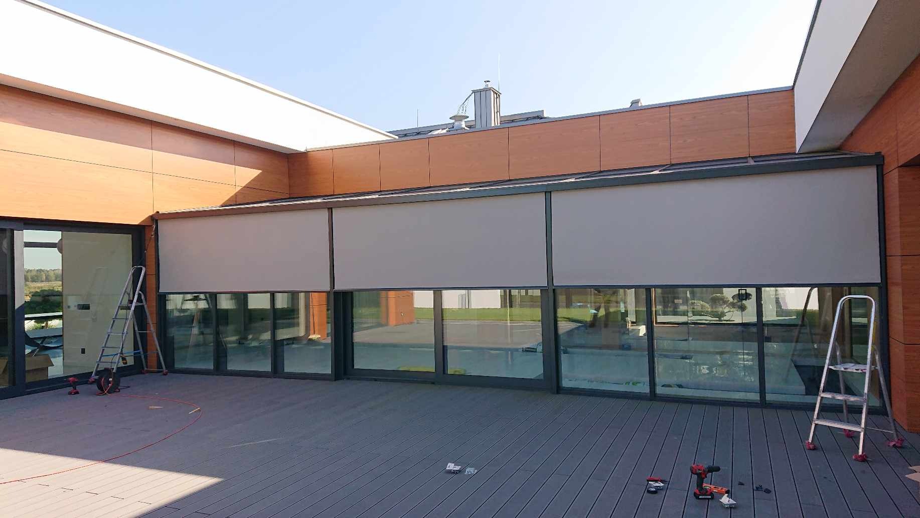 Zewnętrzne osłony okienne w kolorze szarym zamontowane na tarasie domu w Nasielsku, widoczne drabiny i narzędzia podczas montażu.