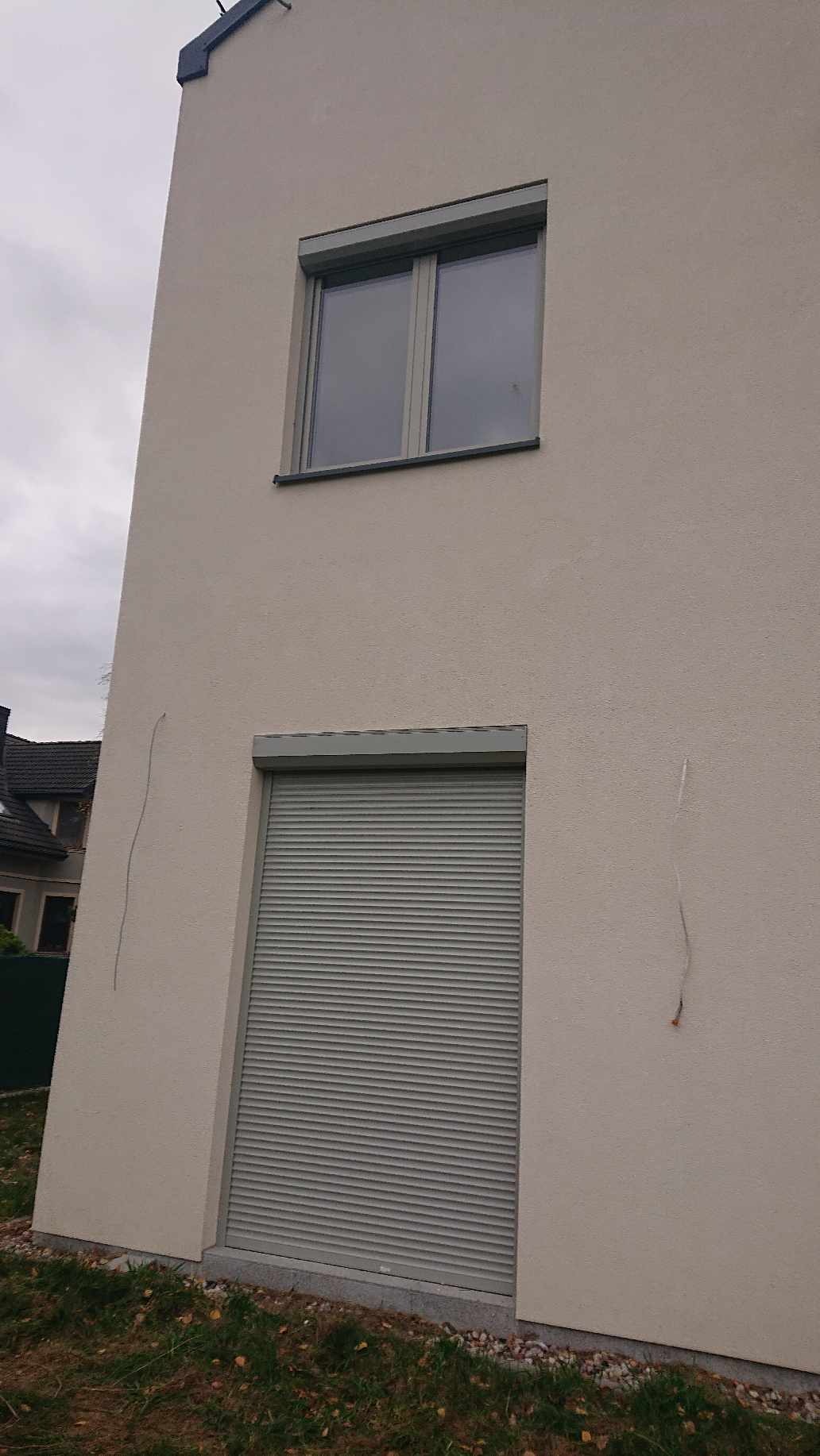 Elewacja budynku z oknem na górze i zamkniętą szarą roletą na dole, widoczne ślady po instalacji na ścianie.