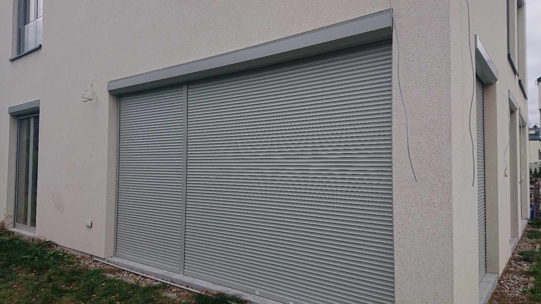 Szare, aluminiowe rolety na oknach i drzwiach balkonowych nowoczesnego domu z jasną elewacją o strukturze baranka, częściowo zwinięte.
