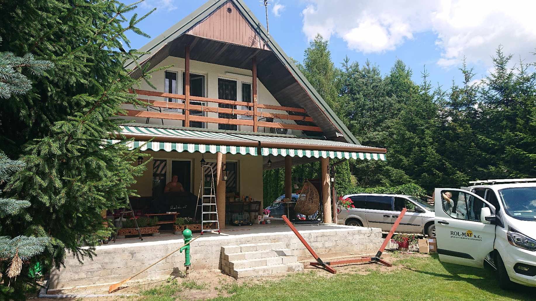 Dom z zamontowaną markizą z zielono-białymi pasami, widoczny samochód dostawczy firmy montującej markizy z otwartymi drzwiami oraz sprzęt montażowy na trawniku.