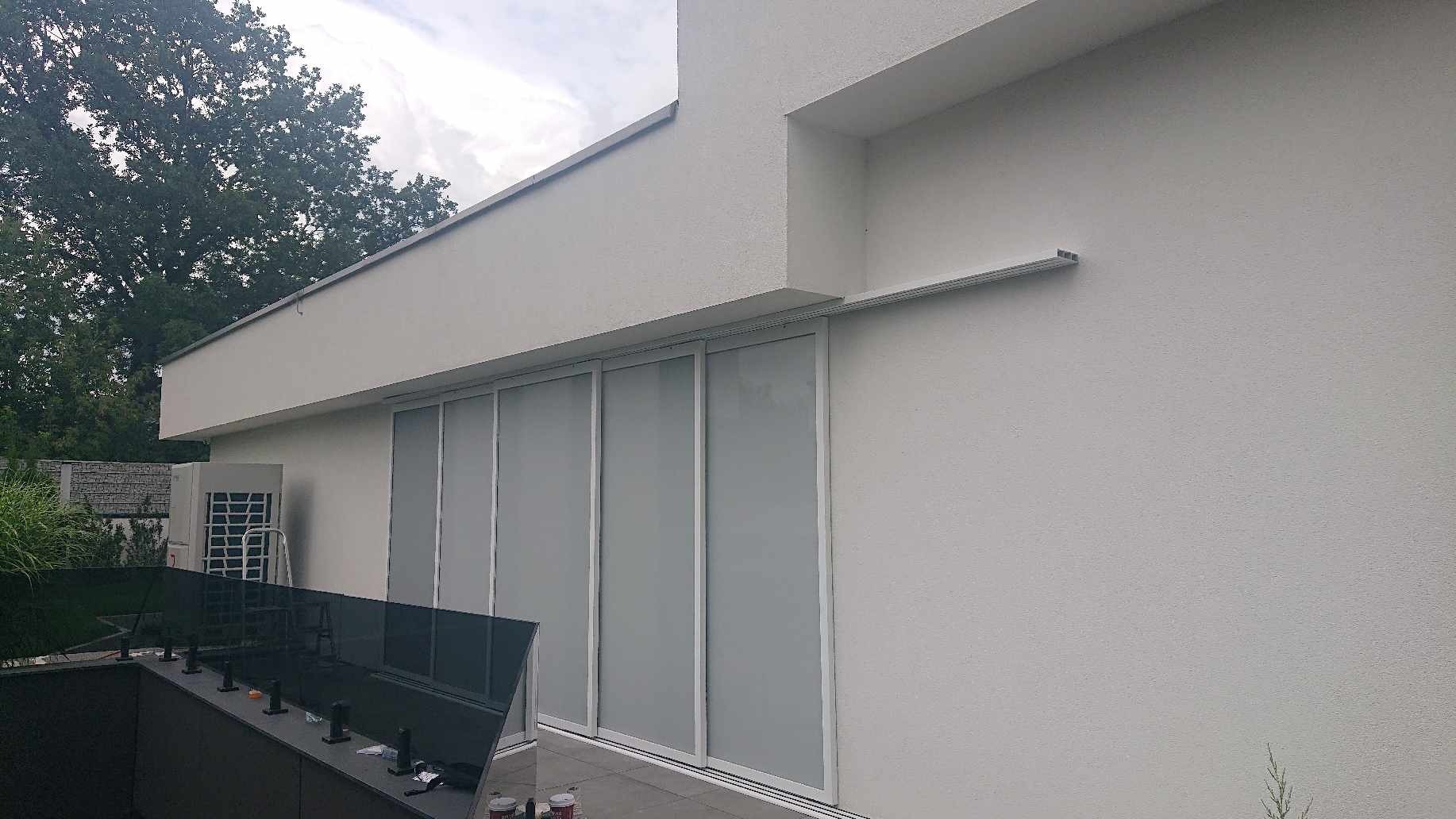 Szare, przesuwne osłony okienne na tarasie nowoczesnego domu z widocznym klimatyzatorem i szklaną balustradą.