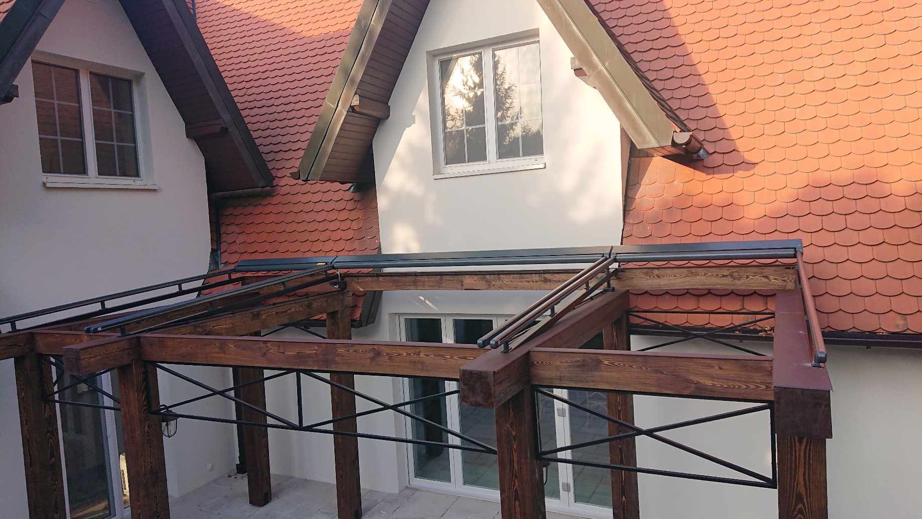 Drewniana pergola z czarnymi metalowymi elementami na tle domu z czerwoną dachówką, widok z góry.