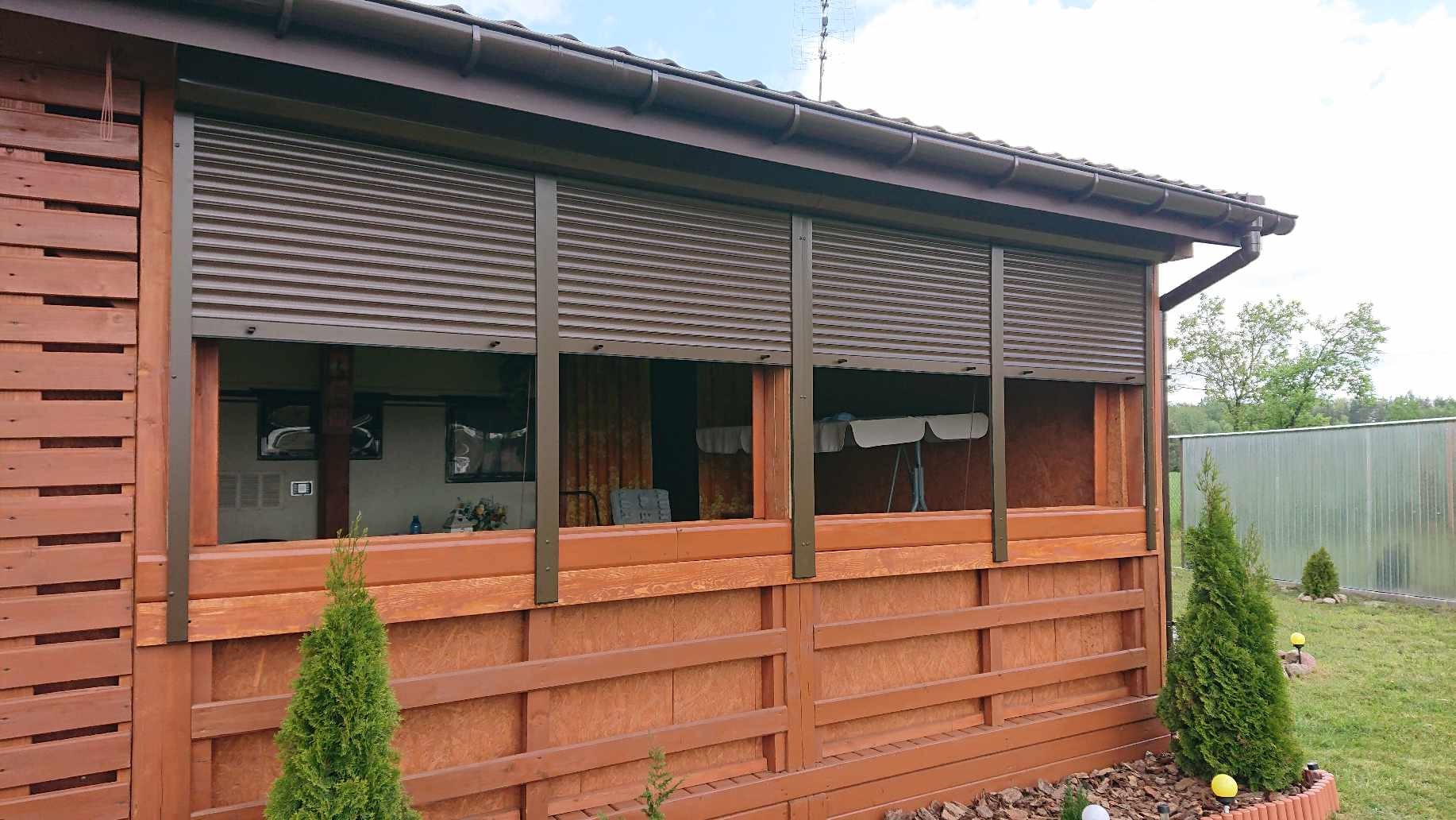 Brązowe, częściowo opuszczone rolety zewnętrzne na drewnianej werandzie, widok z zewnątrz na ogród.