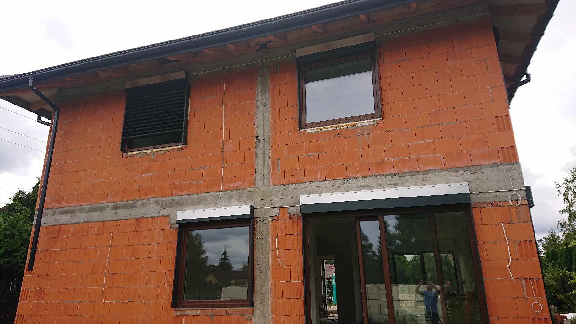 Surowy budynek z czerwonej cegły z zamontowanymi oknami i częściowo zamontowanymi osłonami okiennymi: żaluzjami fasadowymi na górnym oknie po lewej stronie i roletami zewnętrznymi nad oknami...