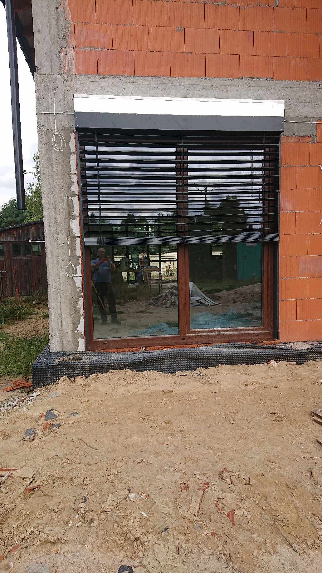 Ciemne poziome osłony okienne zamontowane w oknie budynku w trakcie budowy, widoczne cegły i piasek na zewnątrz.