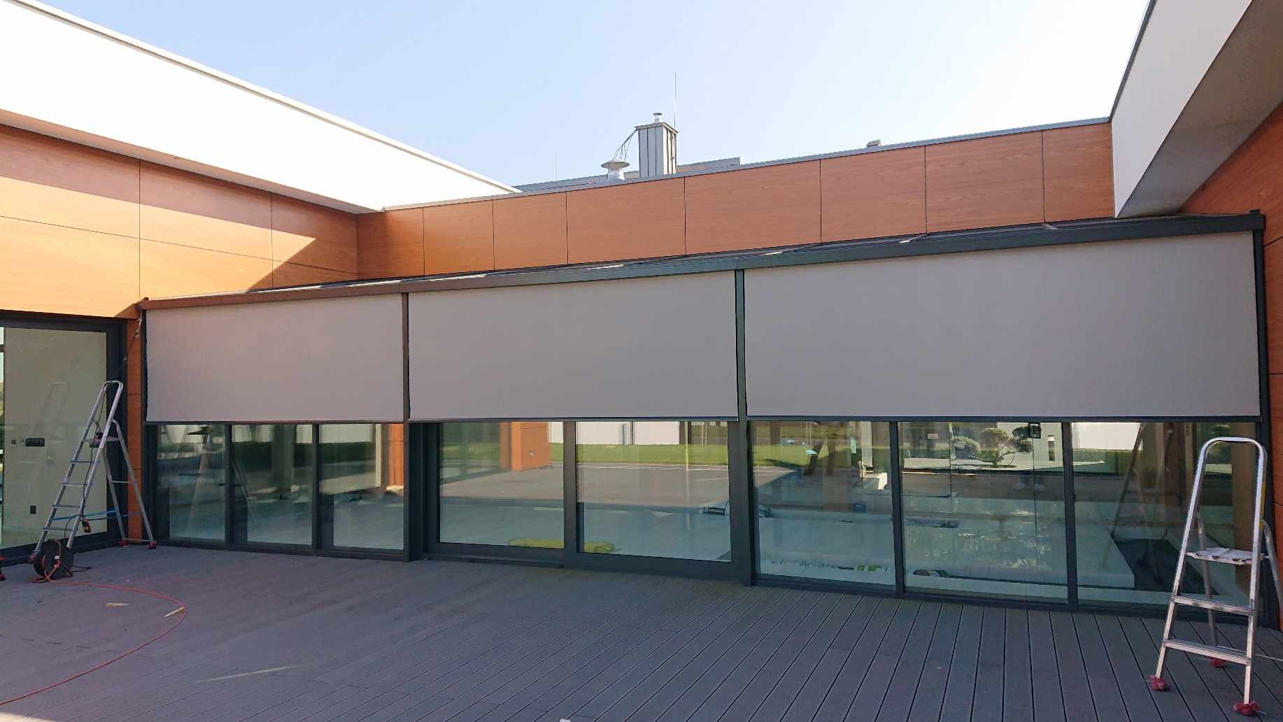 Zewnętrzne osłony okienne w kolorze szarym, zamontowane na nowoczesnym budynku z drewnianą elewacją, widoczne drabiny montażowe po bokach.