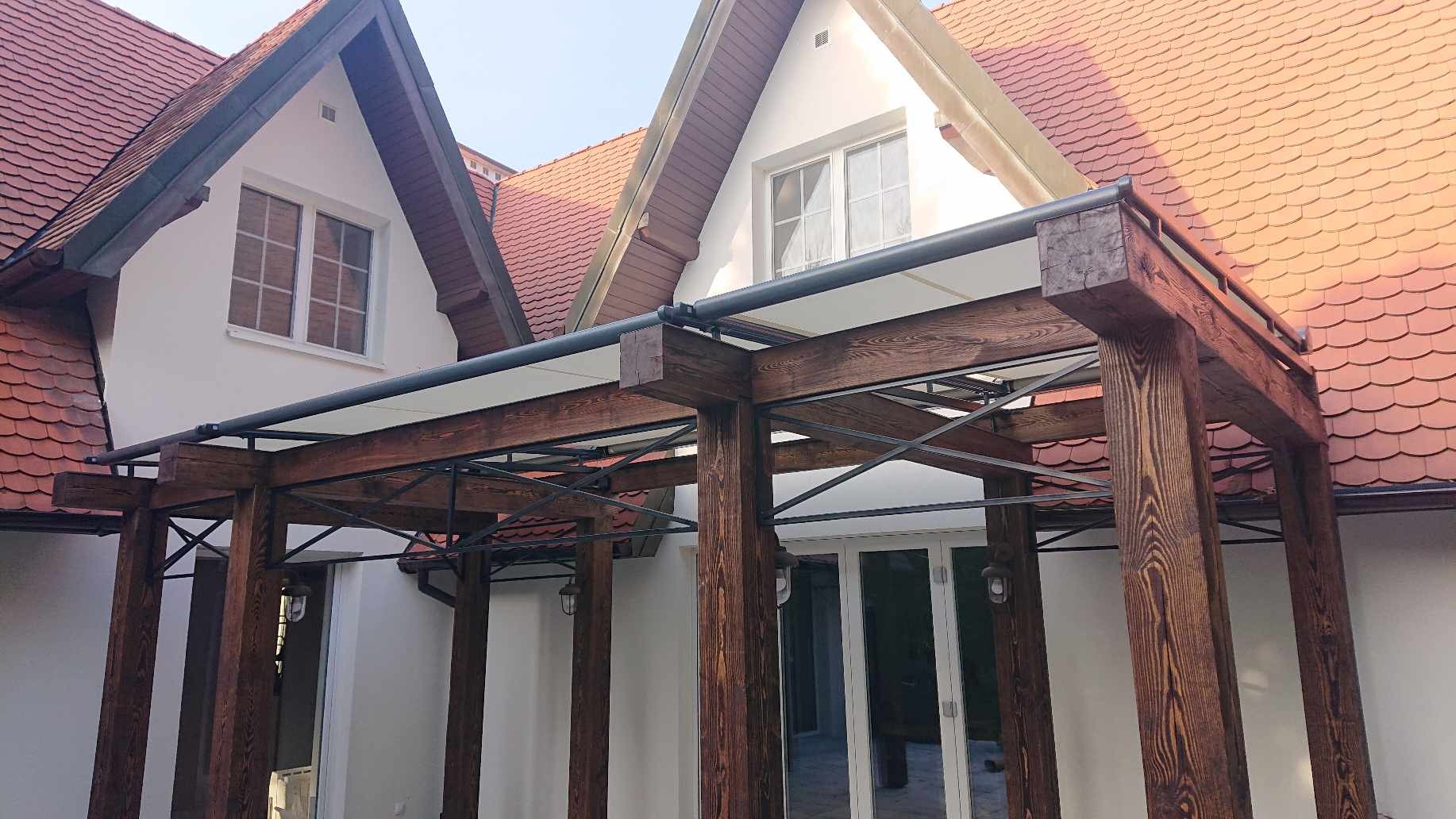 Drewniana pergola z metalowymi wzmocnieniami, przylegająca do białego domu z czerwoną dachówką, widok z dołu.