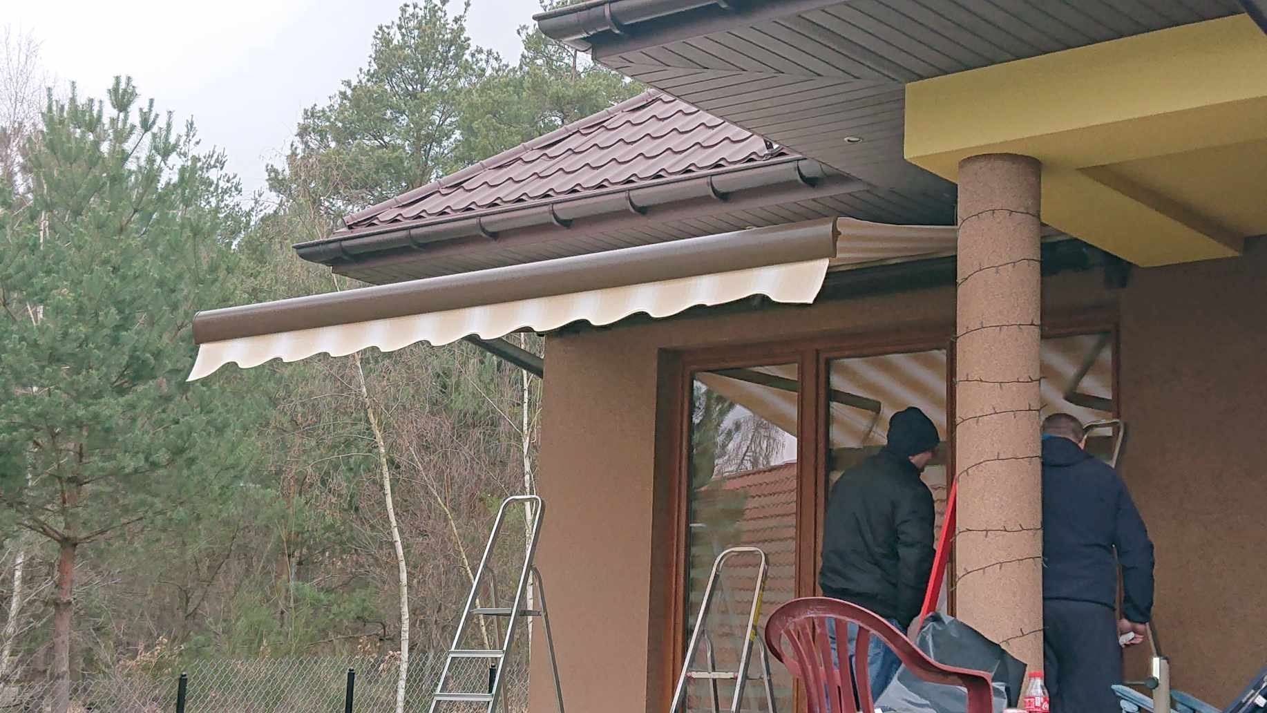 Beżowa markiza z falbaną zamontowana nad oknem domu, widoczne dwie osoby przy pracy, drabiny oparta o ścianę budynku.