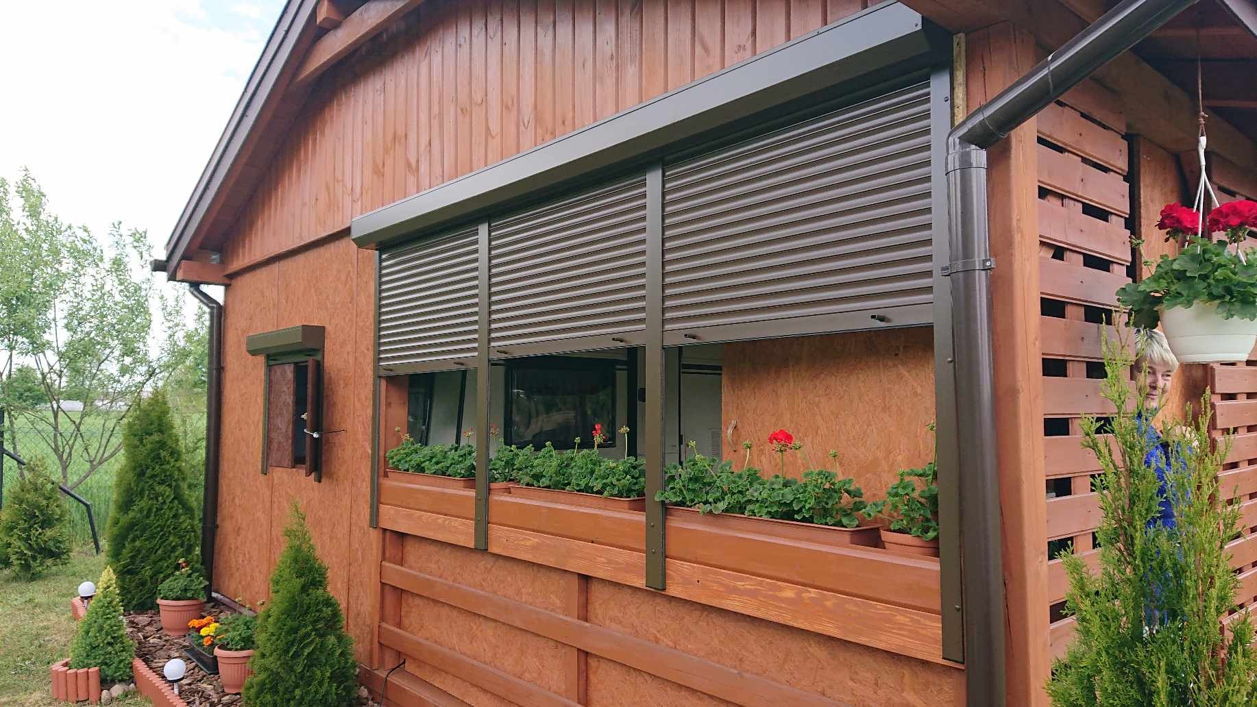 Brązowe rolety zewnętrzne częściowo opuszczone na oknach drewnianego domu z kwiatami w donicach na parapecie, widok z zewnątrz.