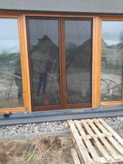 Montaż moskitiery ramkowej w brązowej ramie okiennej, widok z zewnątrz na niedokończony trawnik i paletę drewnianą.