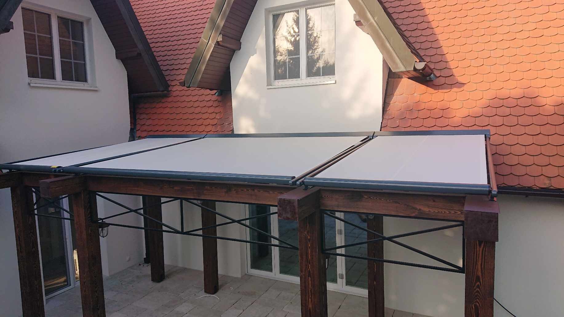 Zadaszenie tarasu z jasną, płócienną osłoną przeciwsłoneczną, wsparte na drewnianej konstrukcji z metalowymi wzmocnieniami, na tle domu z czerwoną dachówką.
