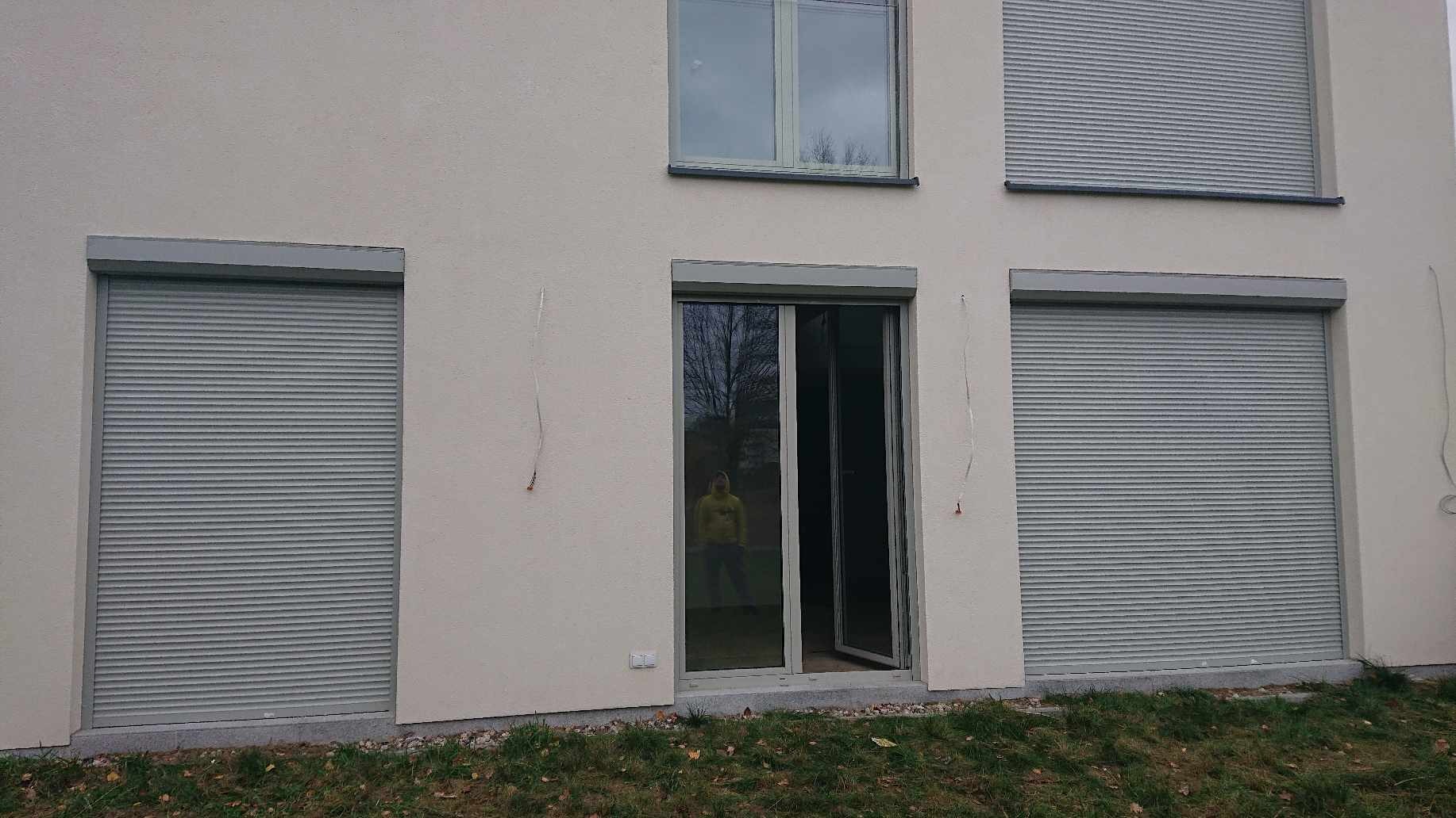 Elewacja budynku z trzema oknami zabezpieczonymi szarymi roletami zewnętrznymi, jedno okno otwarte z odbiciem osoby w środku.