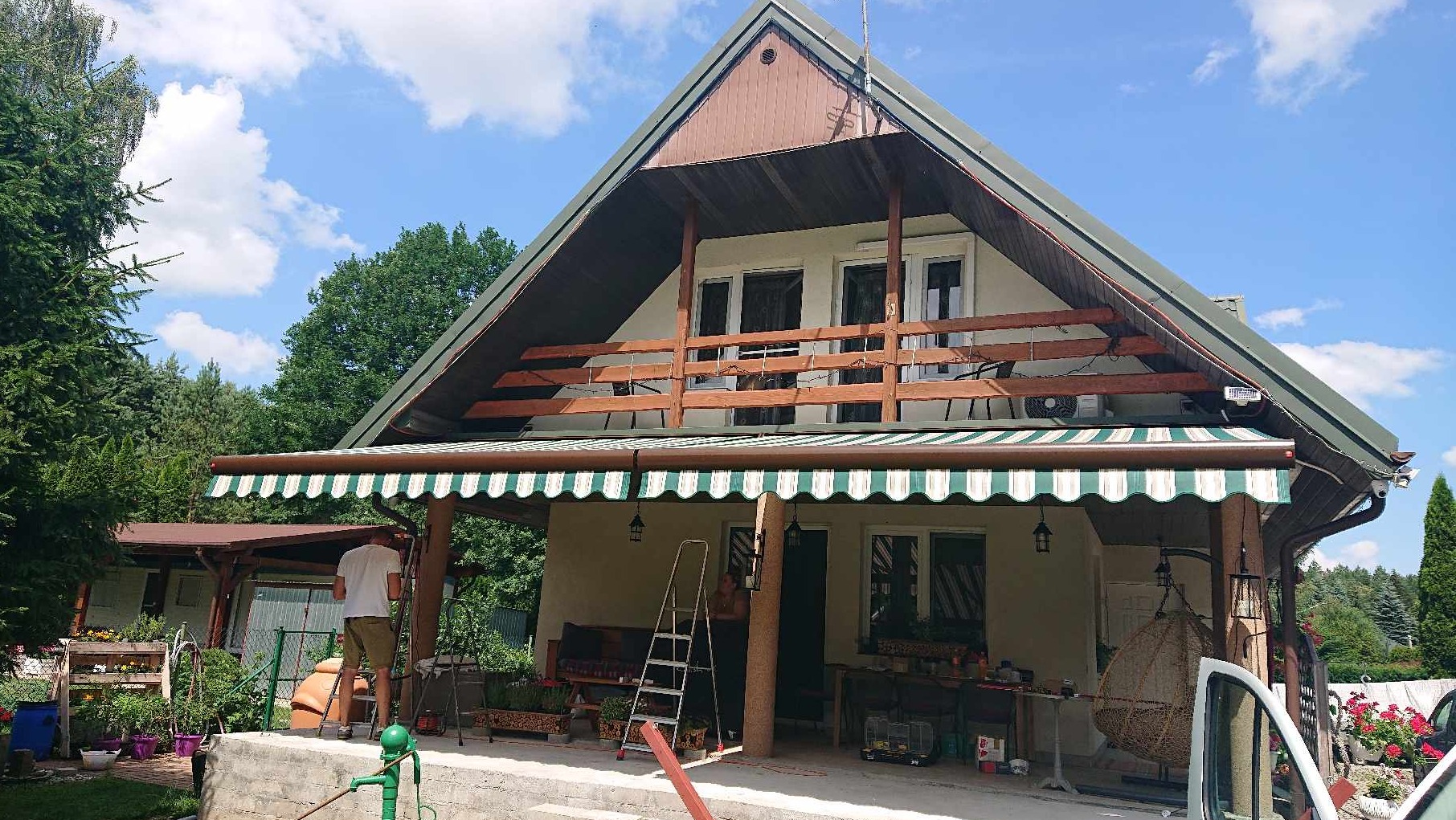 Montaż markizy z zielono-białym pasem na tarasie domu z drewnianym balkonem; pracownik na drabinie, widoczny w tle ogród i błękitne niebo.
