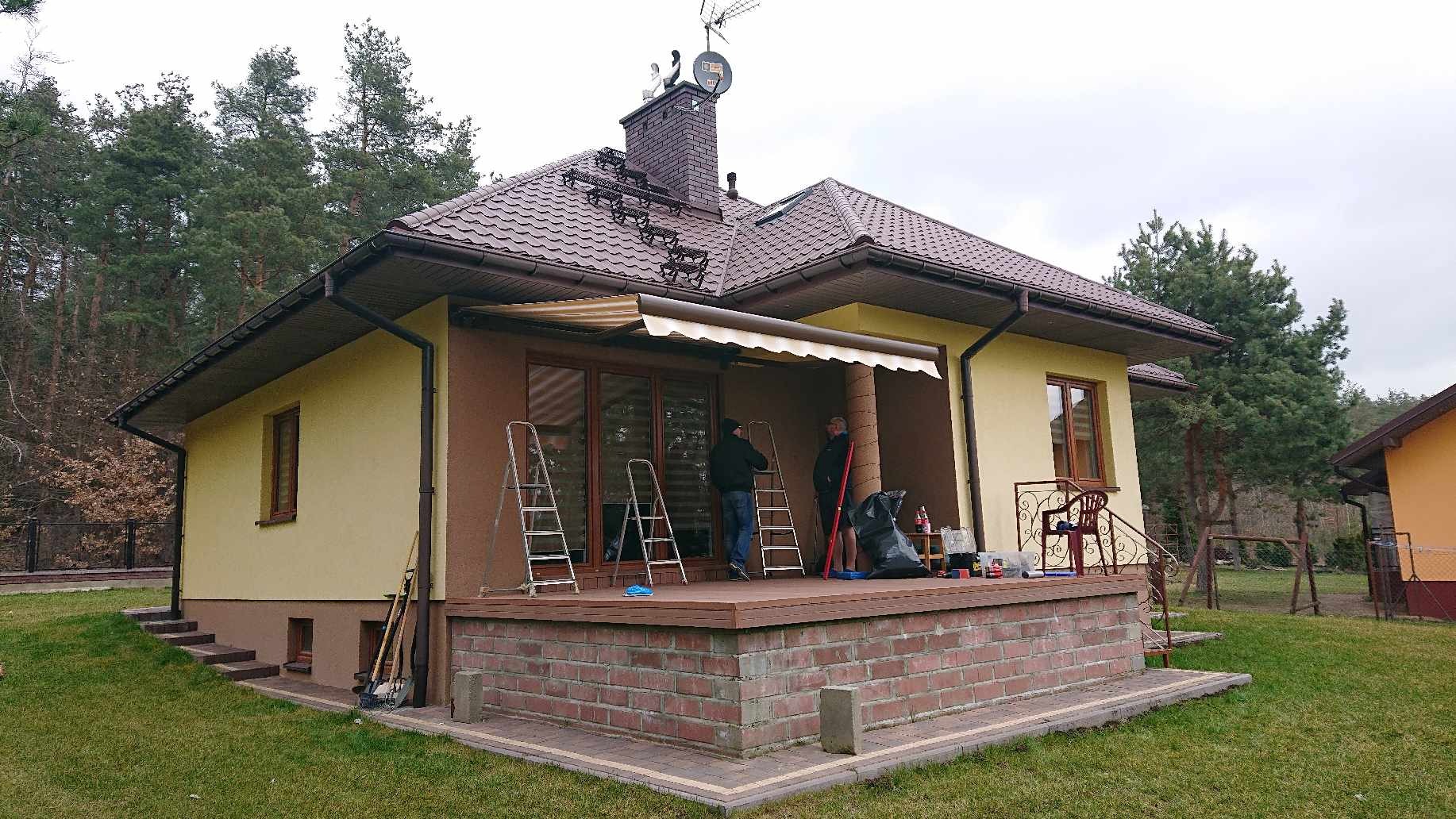 Montaż zwijanej markizy nad tarasem domu jednorodzinnego, widoczne dwie osoby na drabinach, narzędzia i akcesoria montażowe na tarasie wykończonym cegłą.