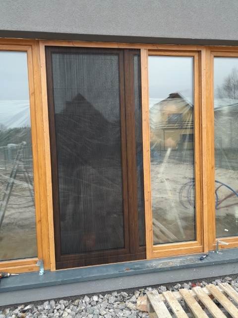 Zamontowana moskitiera ramkowa w drewnianej ramie okiennej, widok z zewnątrz, na tle budynku w budowie.