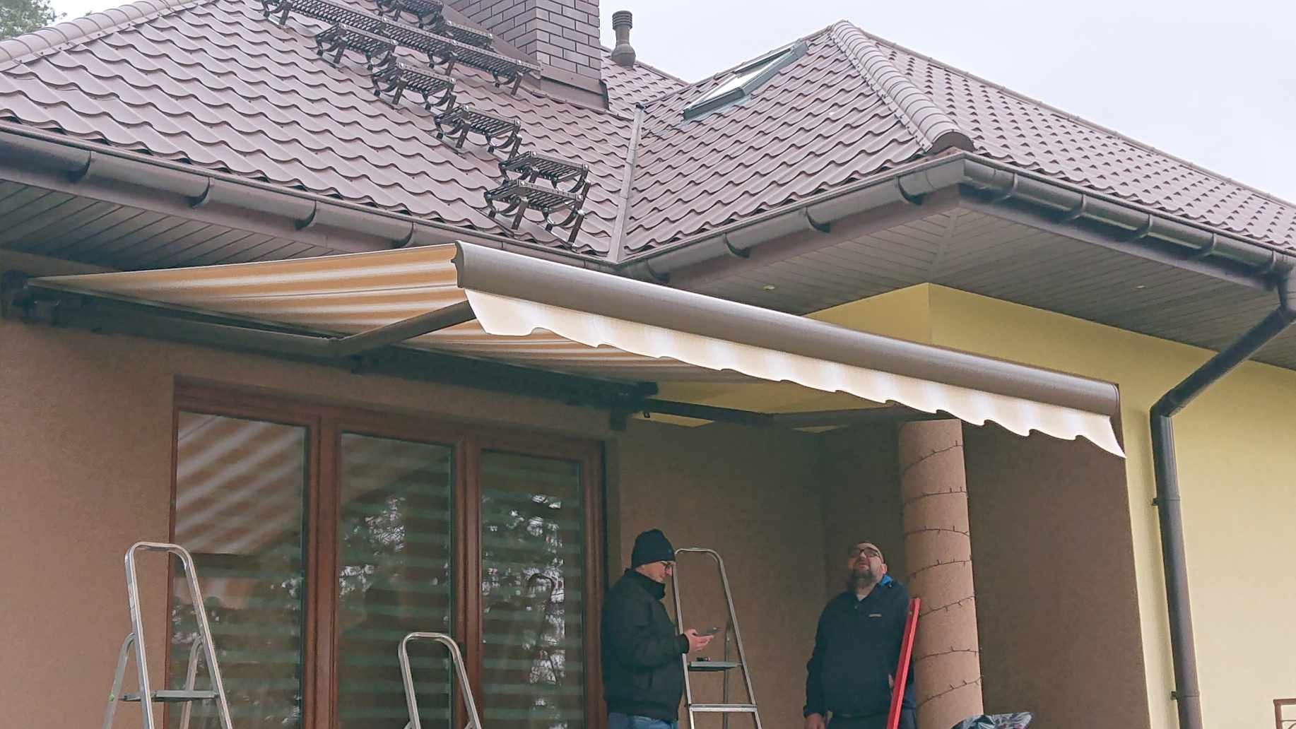 Montaż zewnętrznej osłony przeciwsłonecznej z kremowo-brązowymi pasami na tarasie domu, widoczni dwaj monterzy przy drabinach.