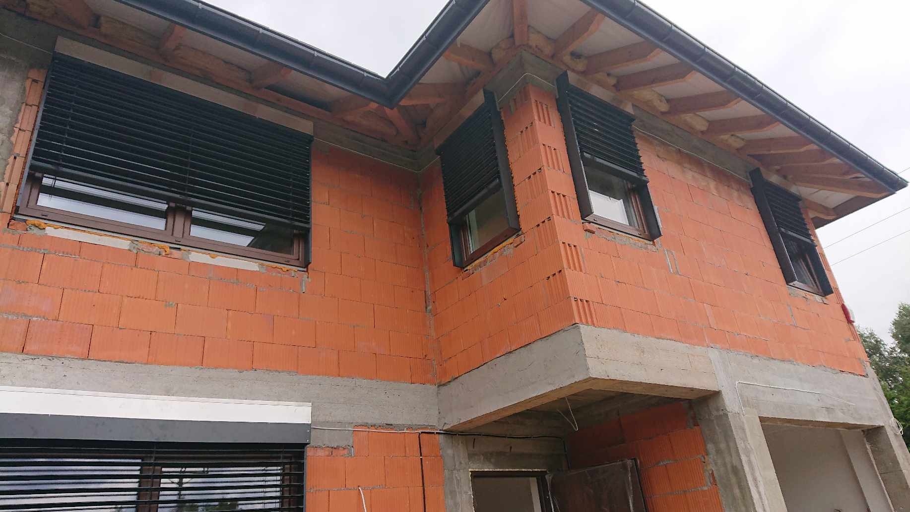 Surowy budynek w trakcie budowy z zamontowanymi czarnymi osłonami okiennymi zewnętrznymi, widoczne cegły, beton i drewniana konstrukcja dachu.