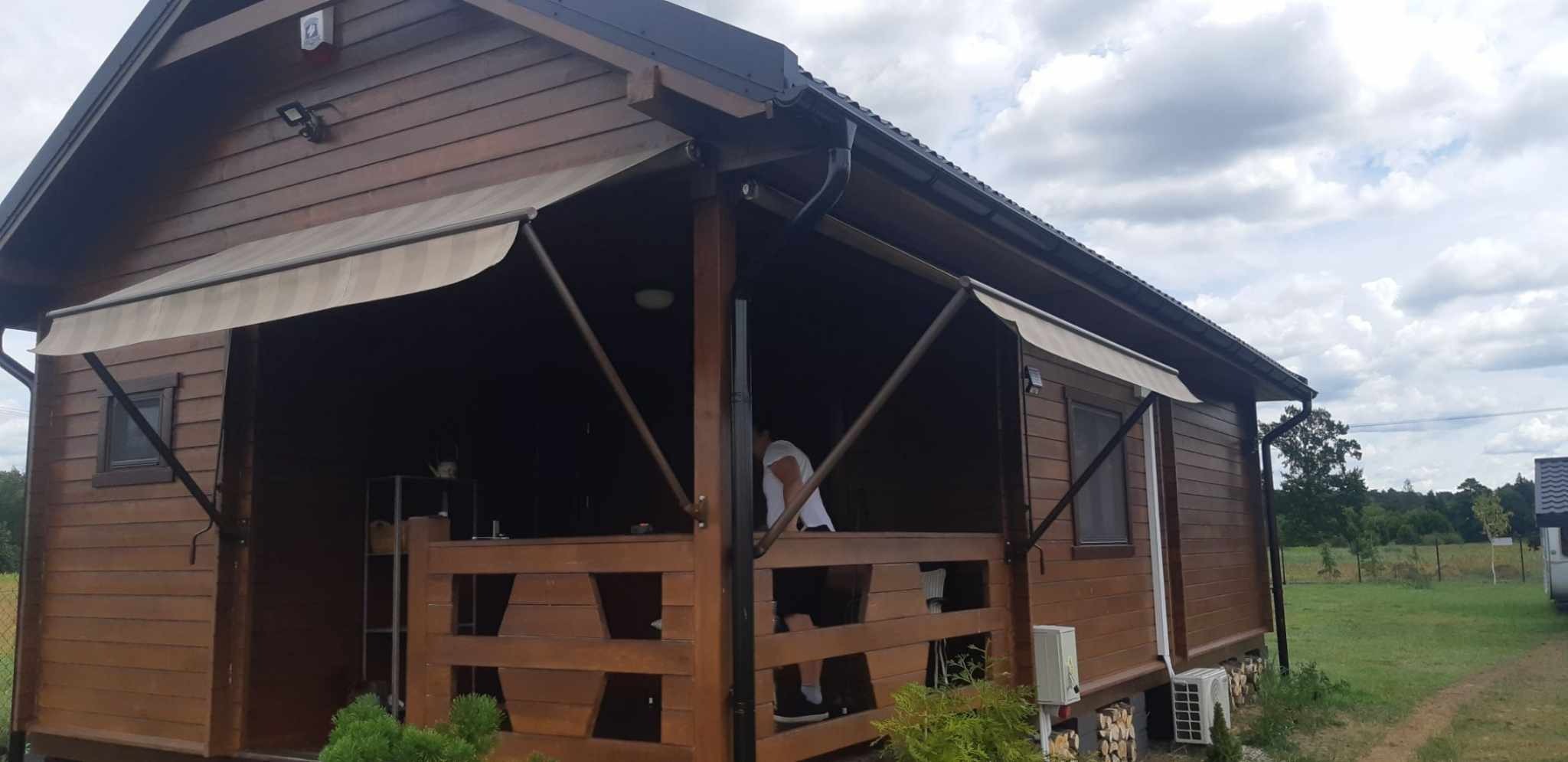 Drewniany domek letniskowy z rozłożonymi markizami przeciwsłonecznymi w beżowe paski, widoczne elementy konstrukcyjne i mechanizm rozkładania, na pierwszym planie drewniana balustrada werandy.