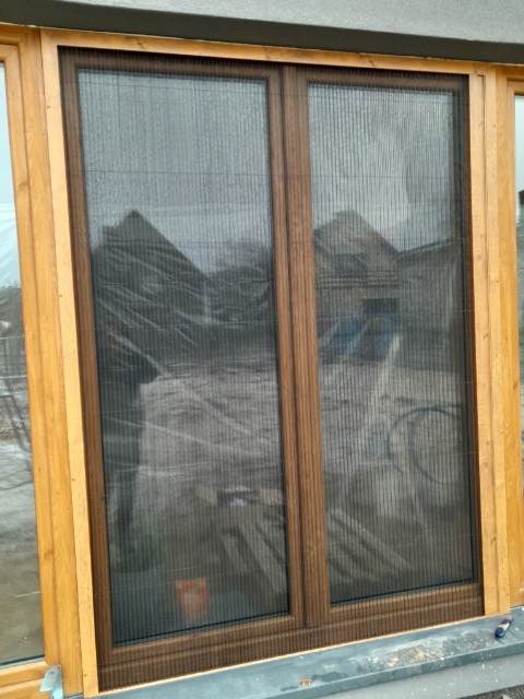 Brązowa rama okienna z zamontowaną siatką przeciw owadom, widok z zewnątrz na budynek w trakcie budowy.