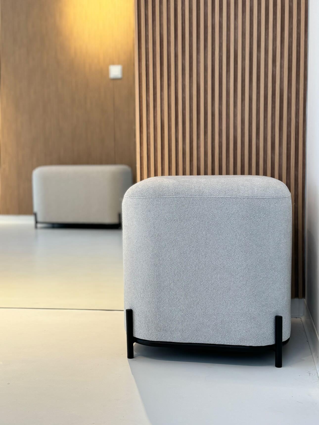 Szary, tapicerowany puf na czarnych, metalowych nóżkach, odbijający się w lustrze na tle drewnianej ściany z pionowych paneli.