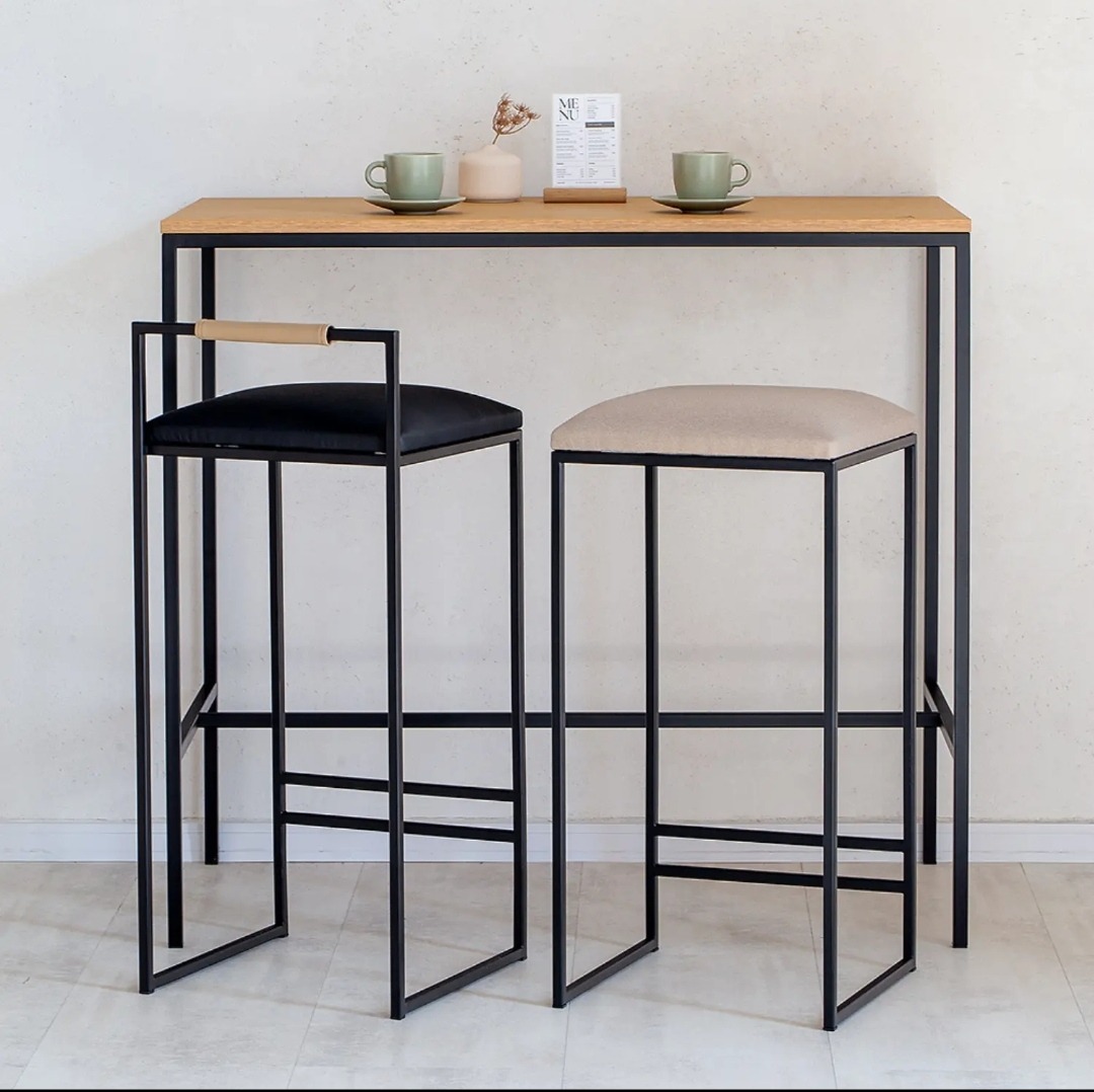 Wysoki stół barowy z blatem z jasnego drewna i czarną metalową ramą, zestawiony z dwoma stołkami barowymi o minimalistycznym designie, jeden z czarnym siedziskiem i oparciem, drugi z beżowym...