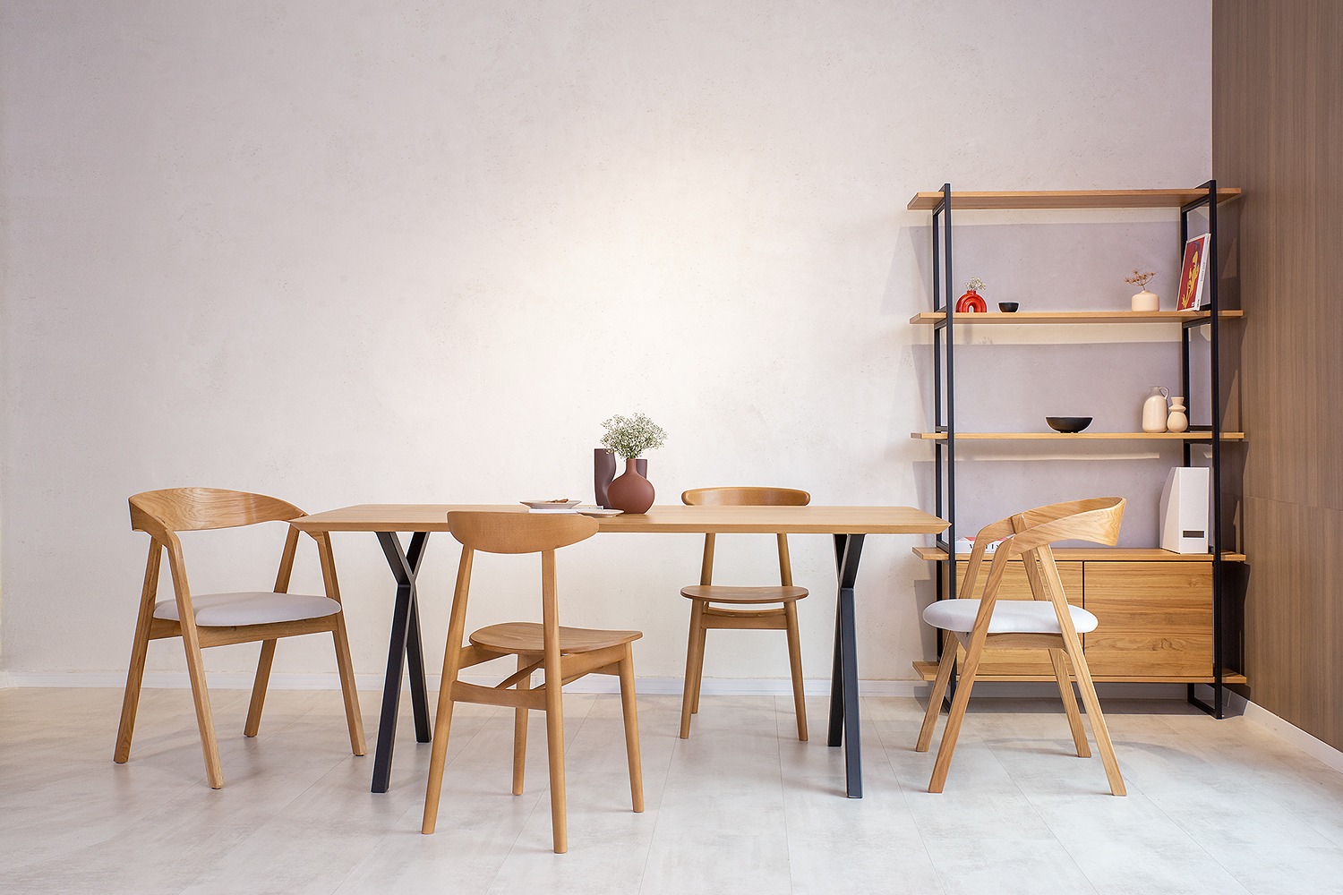 Jasny drewniany stół z czarnymi nogami i czterema krzesłami w minimalistycznym stylu, na blacie wazon z drobnymi białymi kwiatami, w tle regał z drewnianymi półkami i czarną ramą.