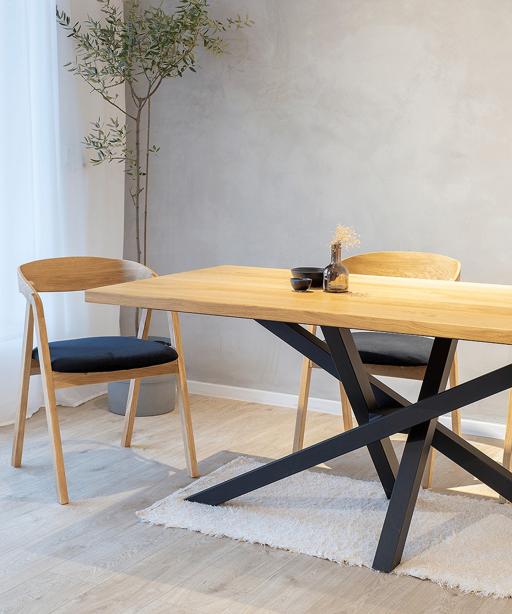 Stół z blatem z jasnego drewna i czarnymi, metalowymi nogami w kształcie litery X, w tle dwie drewniane krzesła z czarnymi poduszkami i roślina w doniczce, na stole dekoracje w postaci miseczek...