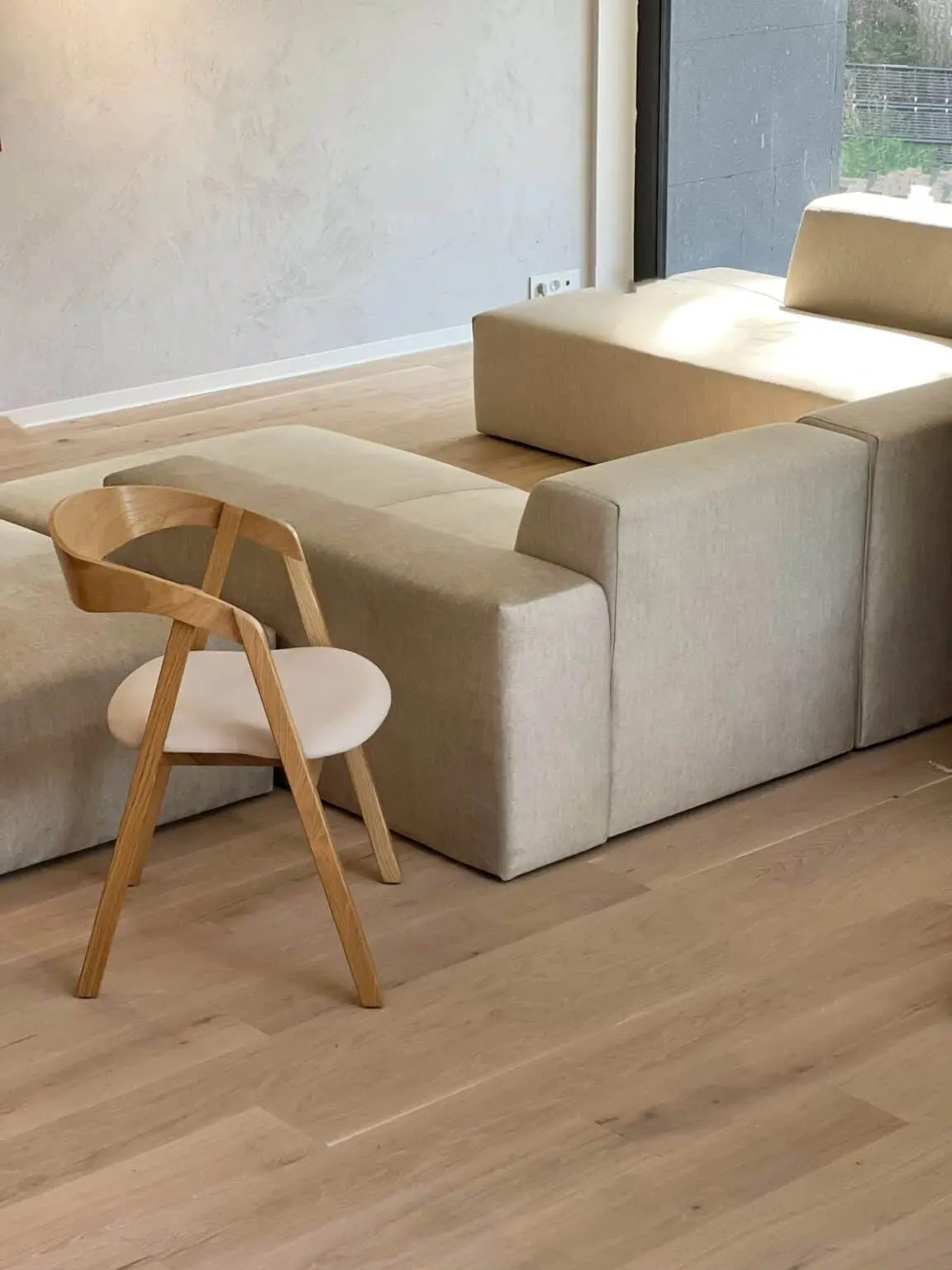 Jasny, modułowy narożnik z tapicerką w odcieniach beżu i krzesło z jasnego drewna z okrągłym, białym siedziskiem na parkiecie. Minimalistyczne wnętrze z dużym oknem.