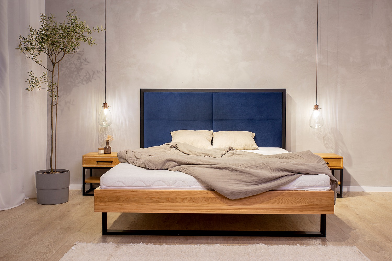 Nowoczesne łóżko z drewnianą ramą i tapicerowanym, granatowym zagłówkiem, oświetlone wiszącymi lampami w stylu Edison, z rośliną doniczkową obok, na jasnej podłodze.