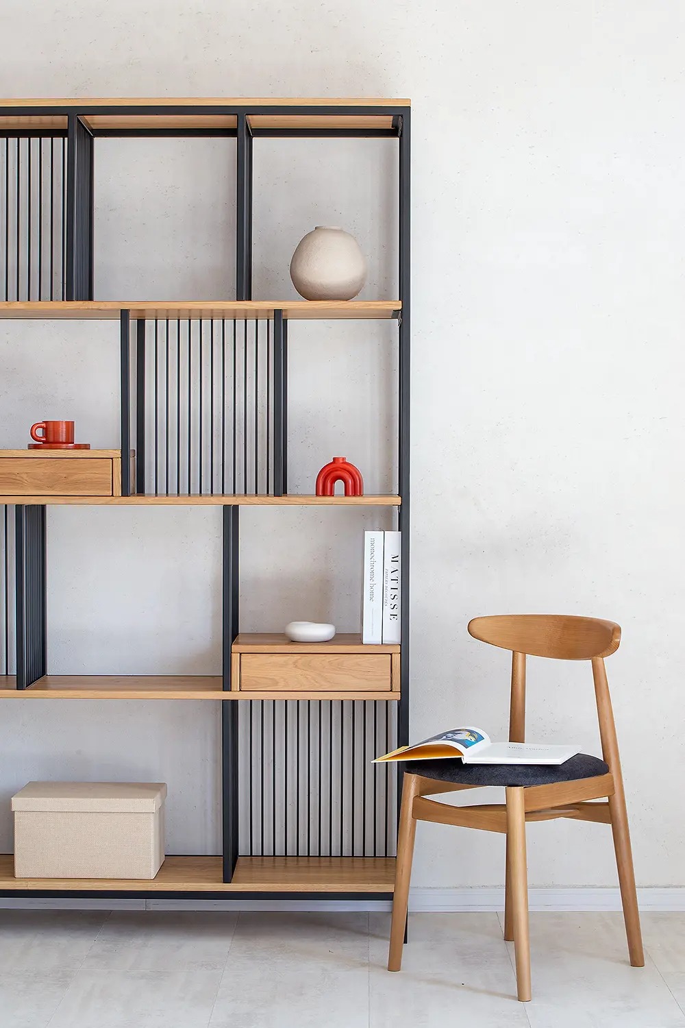 Regał z drewnianymi półkami i czarną metalową ramą, dekorowany ceramiką i książkami, obok drewniane krzesło z otwartą książką.