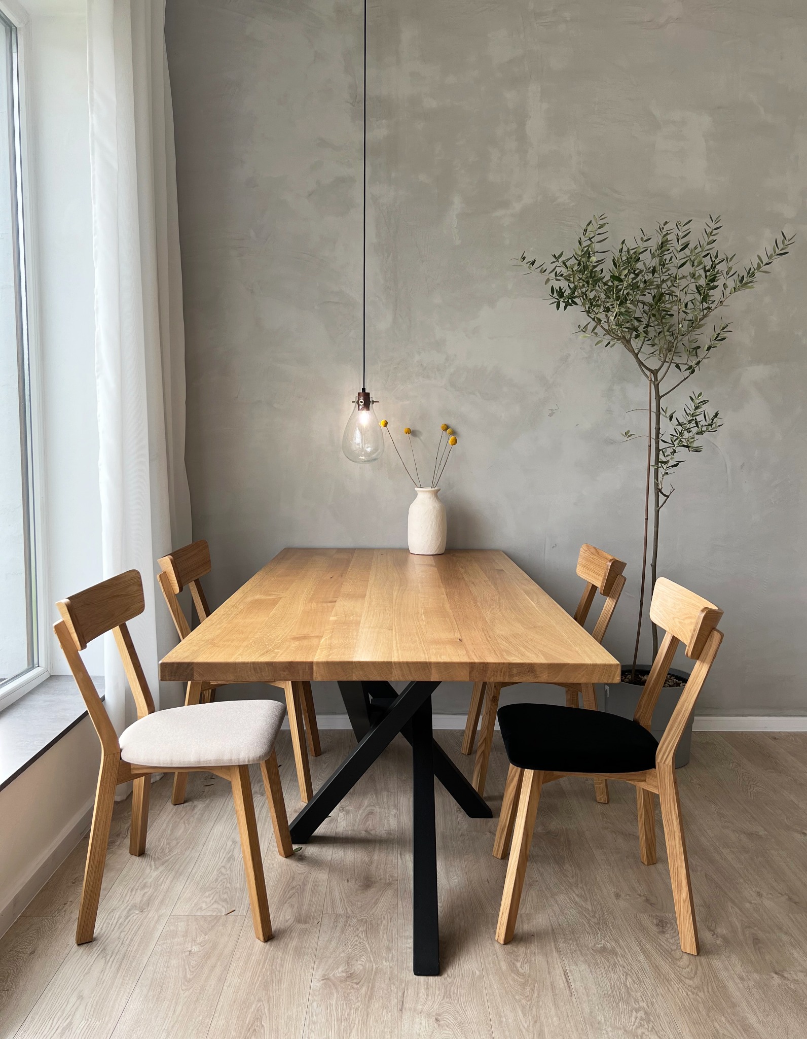 Stół z jasnego drewna z czarną, metalową podstawą w jadalni, otoczony drewnianymi krzesłami z tapicerowanymi siedziskami w kolorze beżowym i czarnym, pod wiszącą lampą w kształcie żarówki i obok...