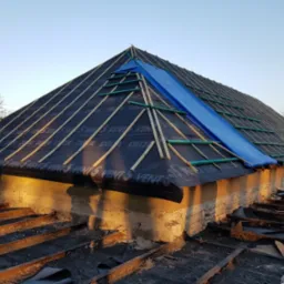 Dach w trakcie prac w Słupsku: czarna membrana dachowa z widocznym nadrukiem, drewniane łaty, fragment niebieskiej plandeki zabezpieczającej przed deszczem, widok z dołu na konstrukcję dachu...