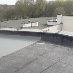 Usługi Dekarskie - Widok z góry na płaski dach budynku pokryty papą termozgrzewalną z widocznymi łączeniami i logo producenta, w tle budynki mieszkalne i drzewa.