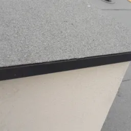 Detale dachu pokrytego papą z czarną listwą wykończeniową, fragment ściany szczytowej o jasnej, chropowatej fakturze.