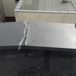 Szare panele blaszane mocowane do dachu pokrytego membraną hydroizolacyjną z widocznym logo producenta, w tle budynek z oknami.