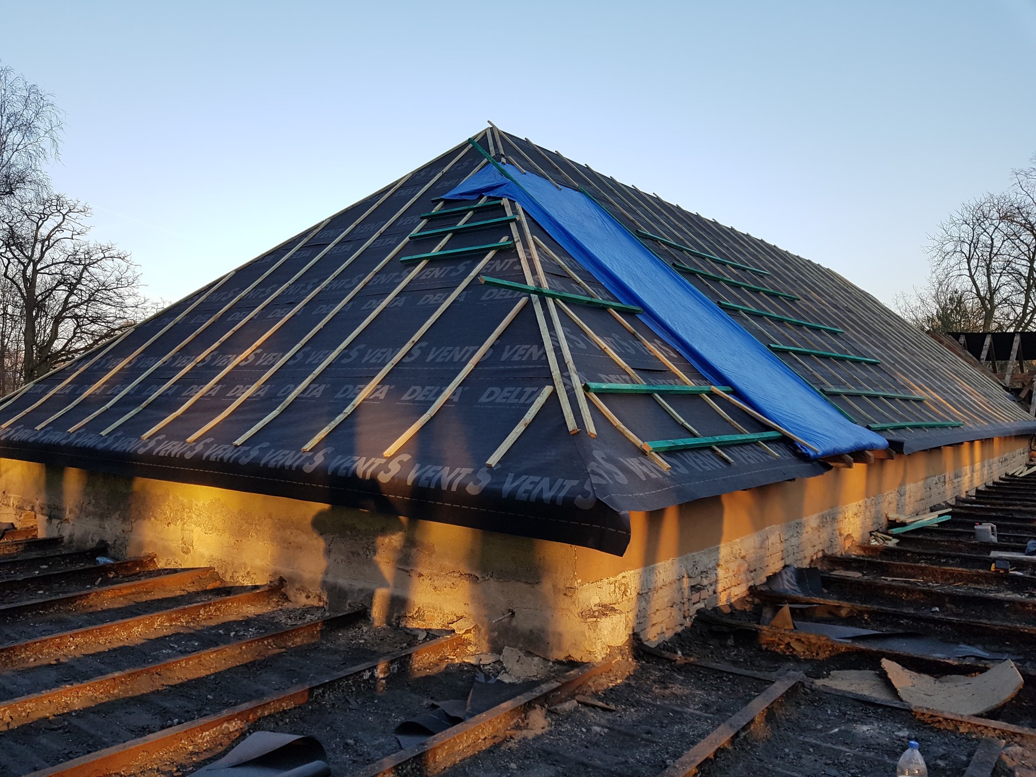 Dach w trakcie prac w Słupsku: czarna membrana dachowa z widocznym nadrukiem, drewniane łaty, fragment niebieskiej plandeki zabezpieczającej przed deszczem, widok z dołu na konstrukcję dachu...