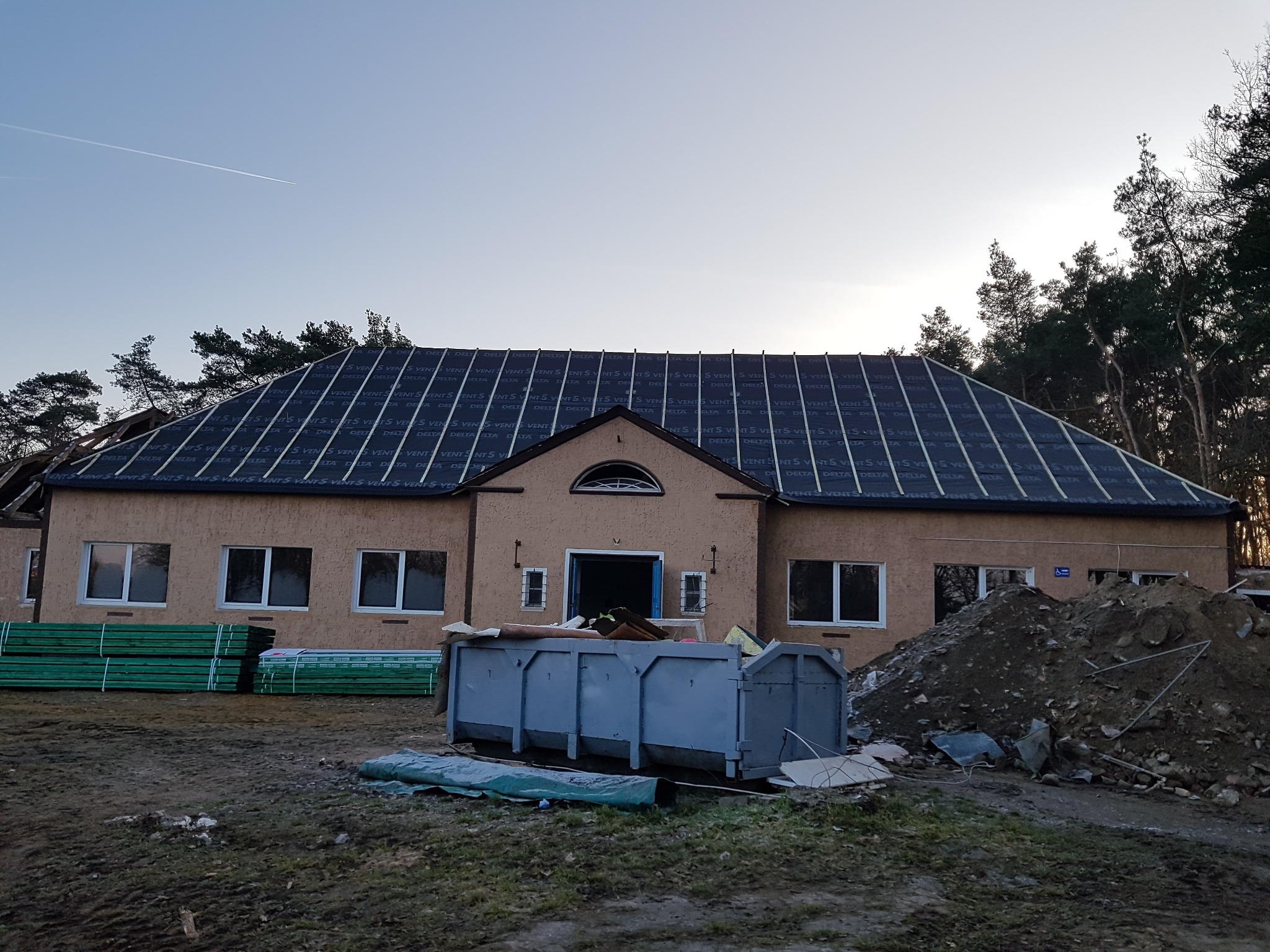 Budynek w trakcie wymiany dachu, widoczna membrana dachowa, stos drewnianych łat, kontener na odpady i hałda ziemi.