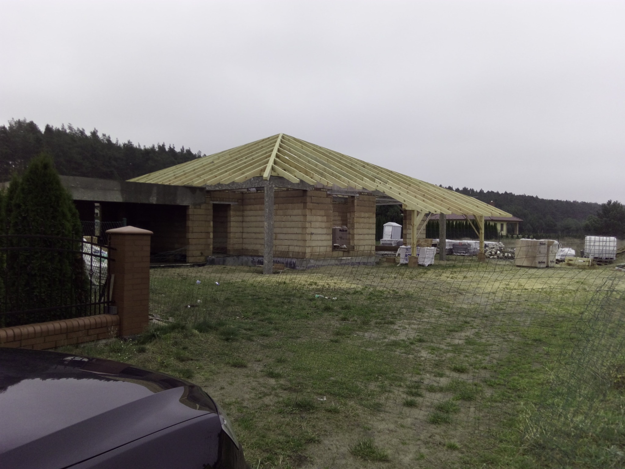 Budowa domu jednorodzinnego w trakcie wznoszenia więźby dachowej, widoczne bloczki konstrukcyjne i przygotowane materiały budowlane na działce.