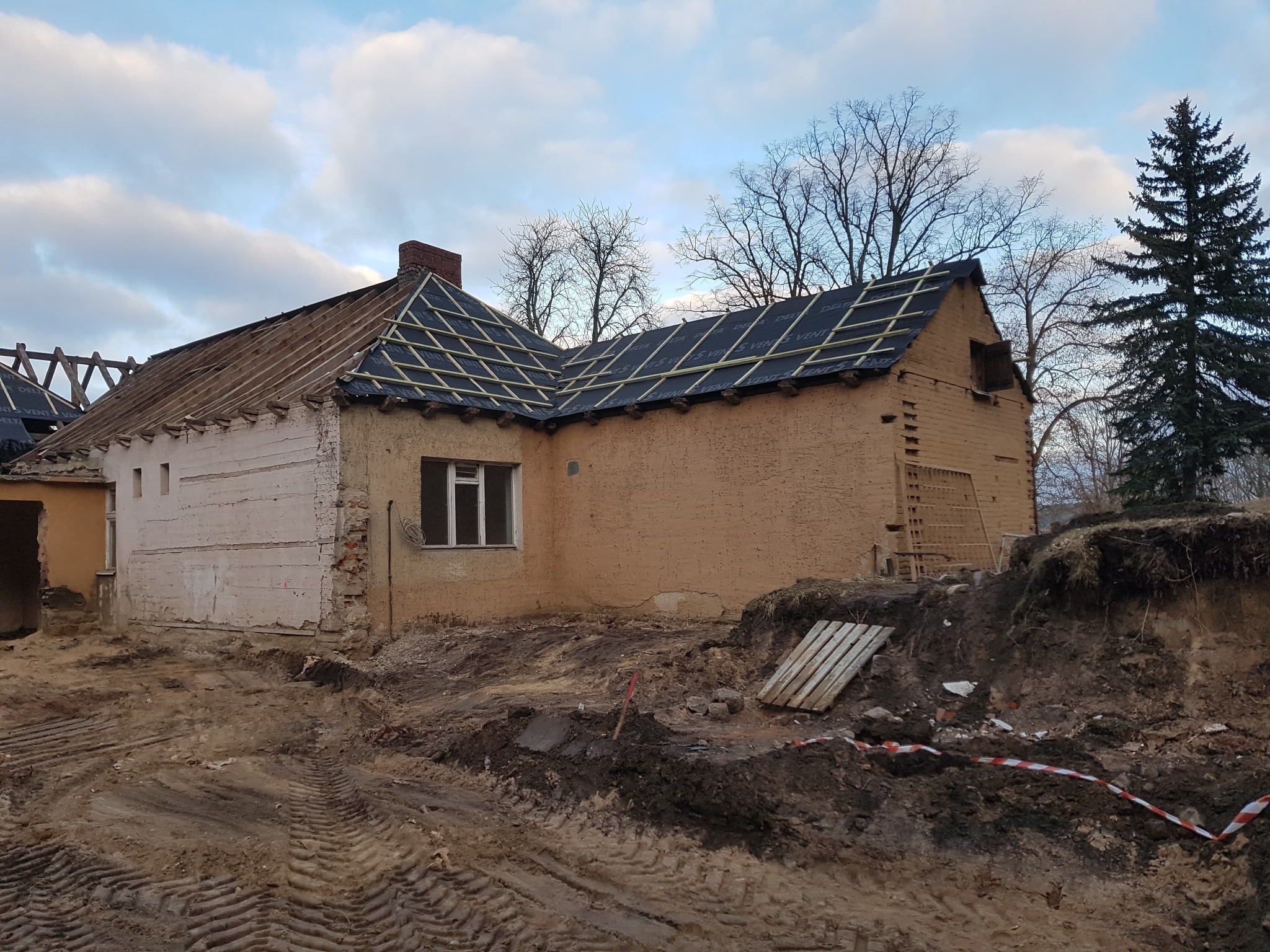 Dom w trakcie remontu dachu: częściowo zdjęte stare pokrycie, widoczne łaty i membrana na nowej części, odsłonięta konstrukcja więźby, obok domu wykopana ziemia i taśma ostrzegawcza.