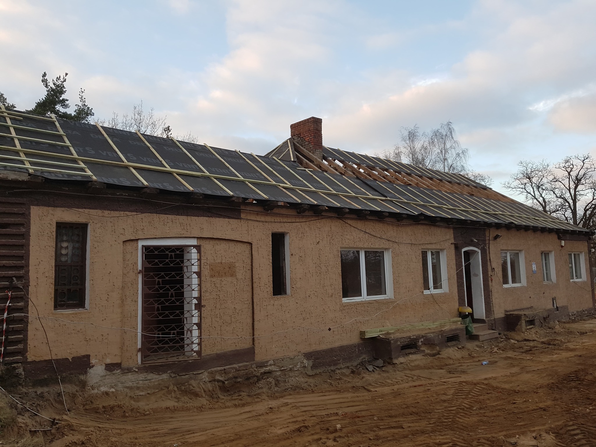 Stary dom z częściowo zdjętym dachem, widoczna membrana dachowa i drewniana konstrukcja łat, ceglany komin w tle, teren wokół rozkopany.